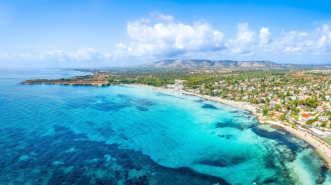 Vista aerea di un mare turchese che incontra una spiaggia sabbiosa con una cittadina costiera, colline verdi sullo sfondo e un cielo azzurro.