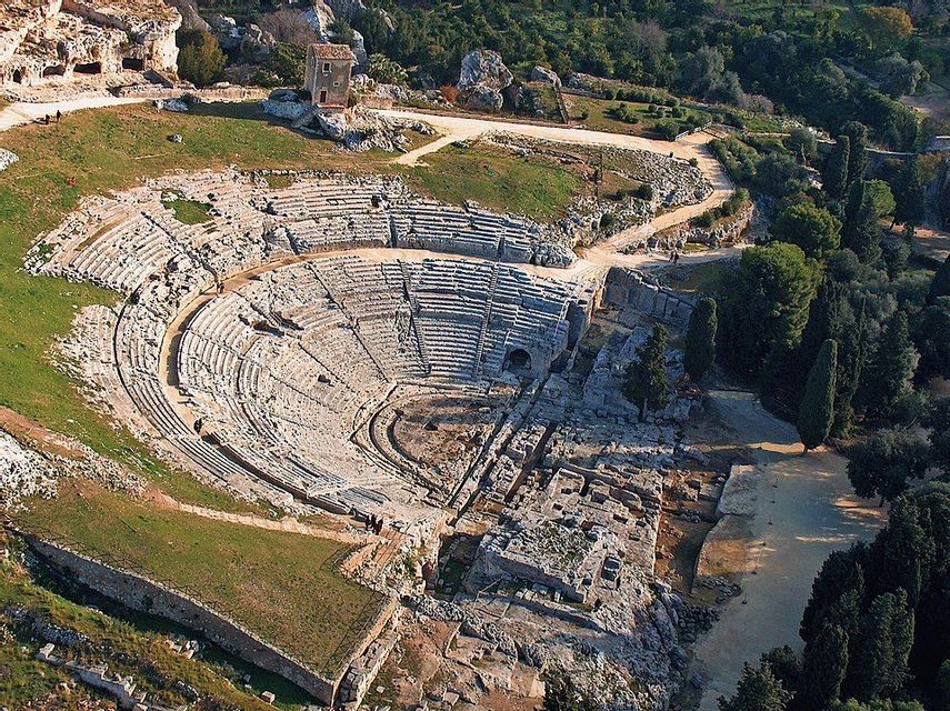 Veduta aerea di un antico anfiteatro in stile romano scavato in una collina rocciosa e circondato da alberi verdi.