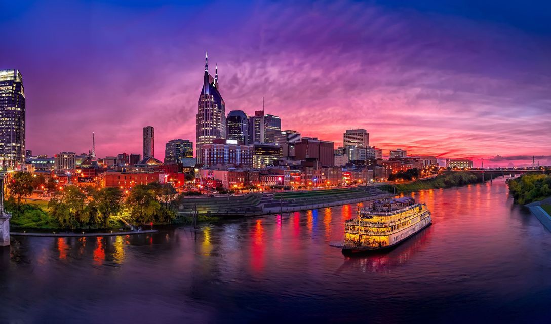 An illuminated riverboat floats on a wide river in front of a modern city skyline under a vibrant purple and pink sky at twilight.