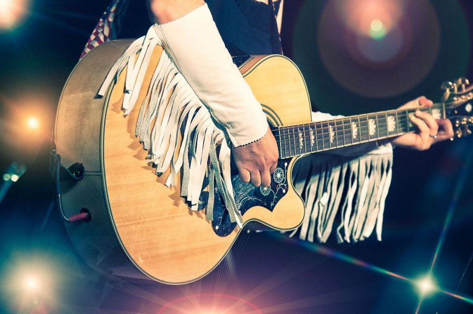 A close-up of a person wearing a white fringed sleeve playing an acoustic guitar on a stage with lens flares.