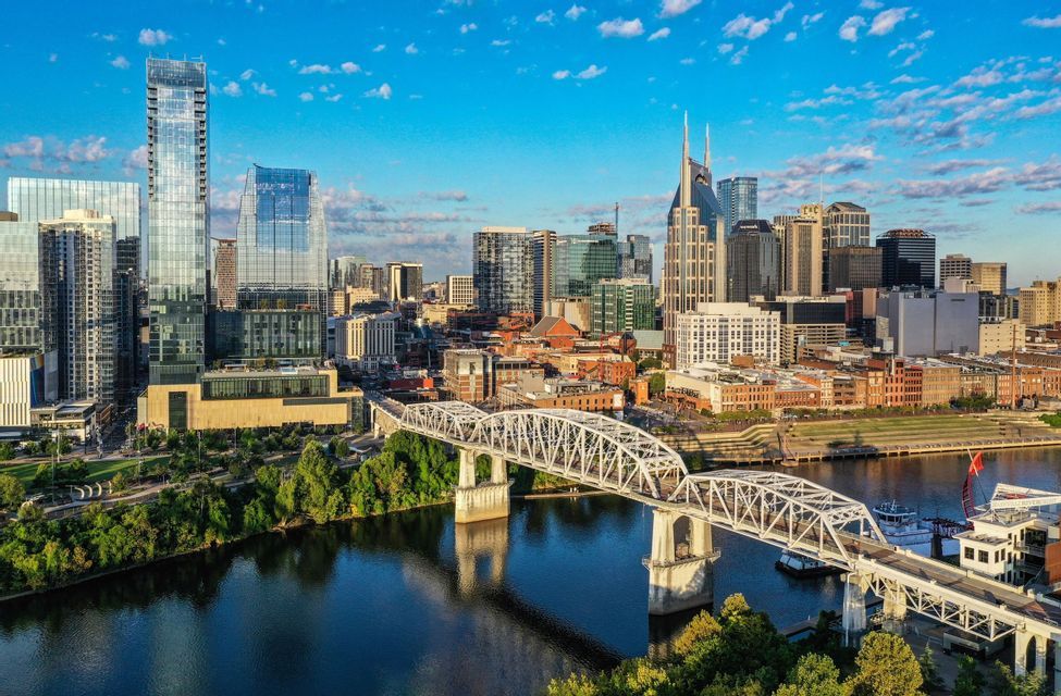 An aerial view of a modern city skyline with skyscrapers and a white truss bridge crossing a river under a blue sky.