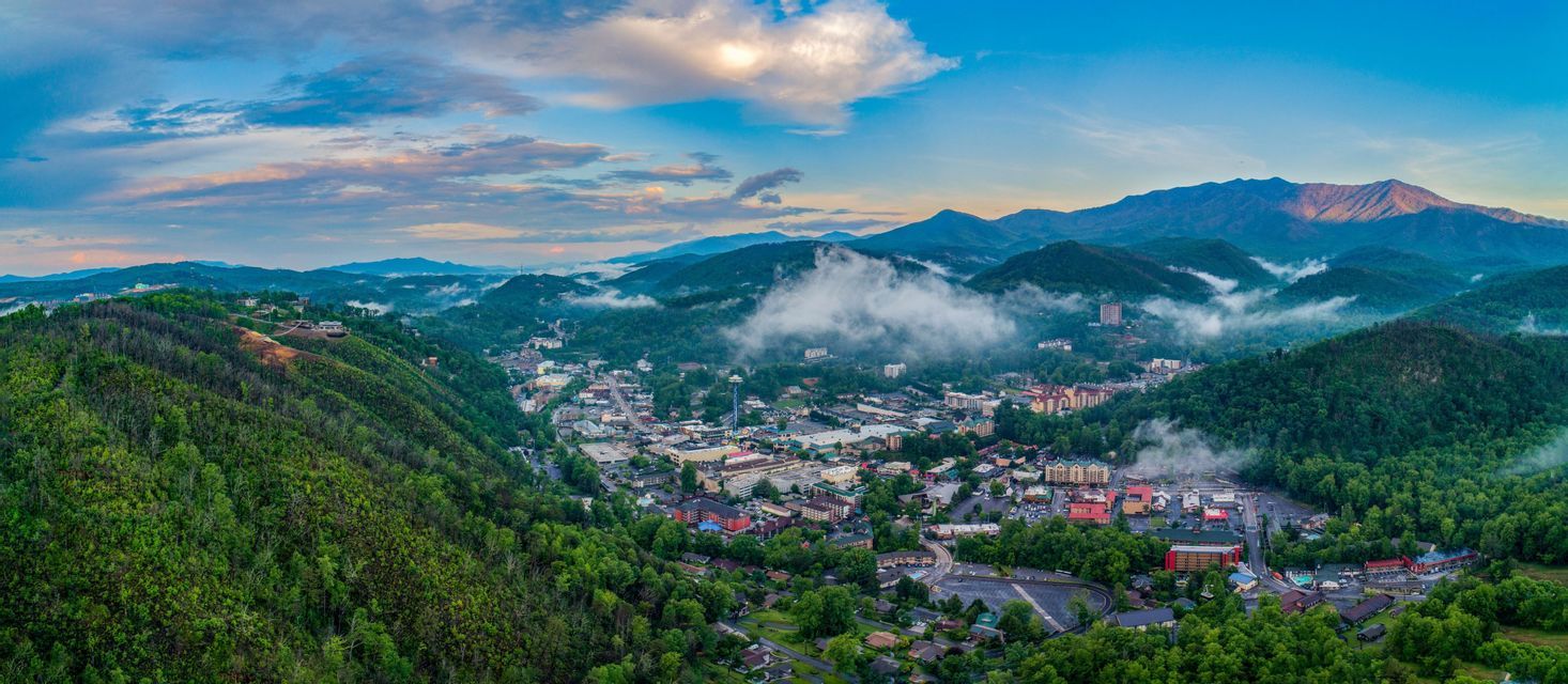 A panoramic aerial view of a town nestled in a green mountain valley with low clouds and mist under a blue sky.