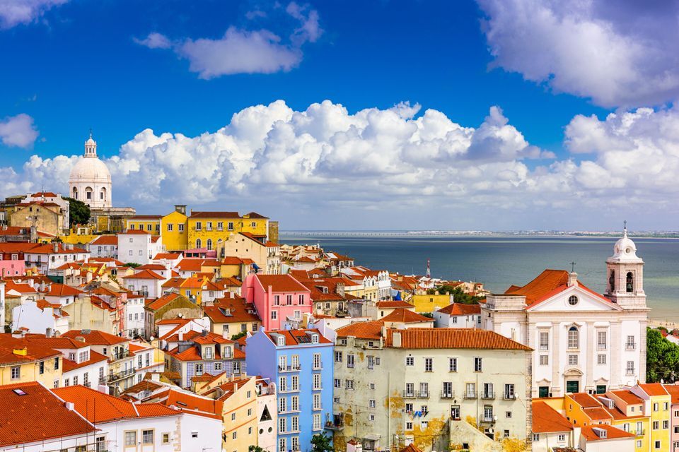 A colorful city with terracotta-roofed buildings on a hillside overlooking a wide body of water under a partly cloudy blue sky.