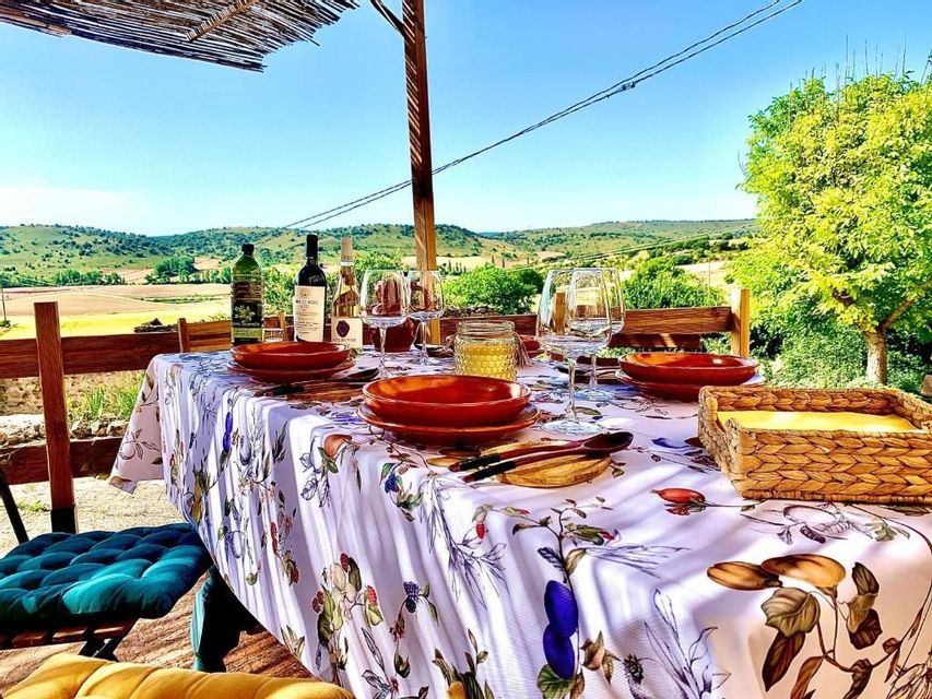 A dining table with a floral tablecloth is set with red plates and wine glasses on a terrace overlooking rolling green hills.