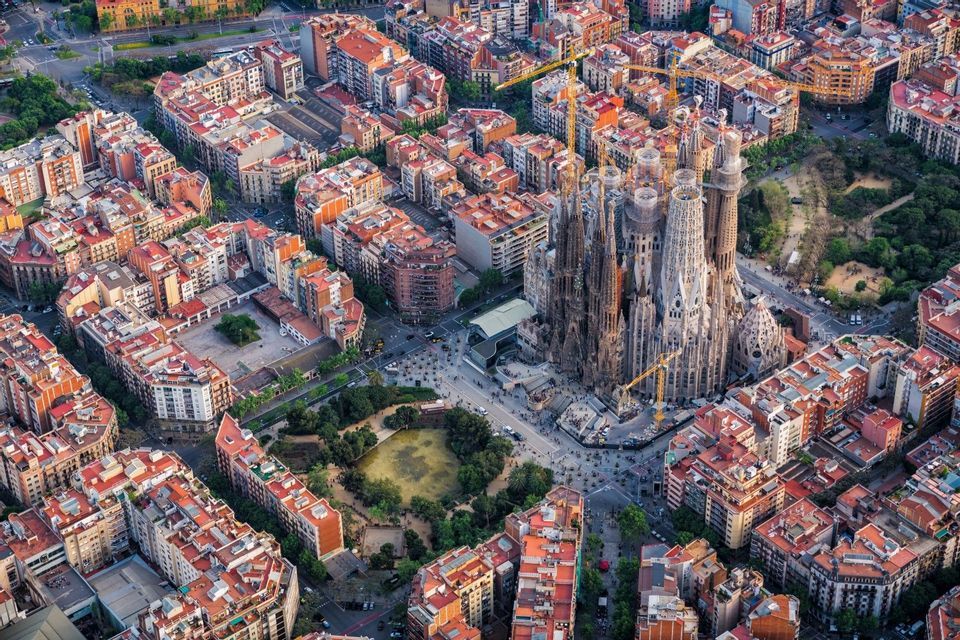An aerial view of a large, ornate cathedral under construction, surrounded by a dense grid of city buildings with red roofs.