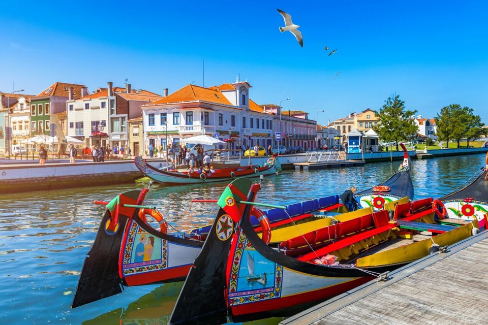 Colorful traditional boats are moored in a city canal, with waterfront buildings and people in the background under a clear blue sky.