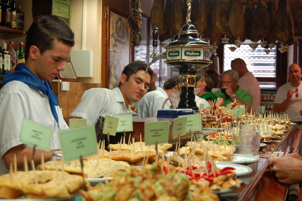 A long wooden counter in a bustling bar, laden with a wide variety of tapas and pintxos for customers.