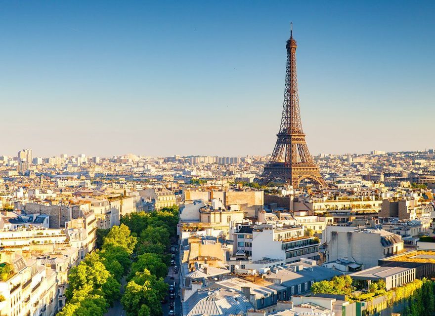 An aerial view of the Paris cityscape with the Eiffel Tower rising above the rooftops under a clear blue sky.
