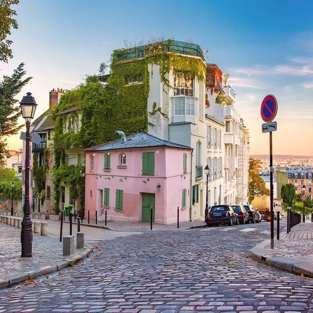 A pink house with green shutters sits on a cobblestone street corner next to an ivy-covered building at sunset.