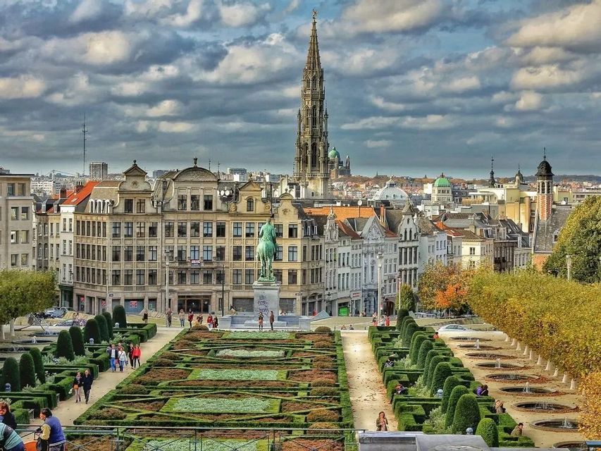 An elevated view of a formal garden with manicured hedges and pathways, leading towards a historic cityscape with a tall spire under a cloudy sky.