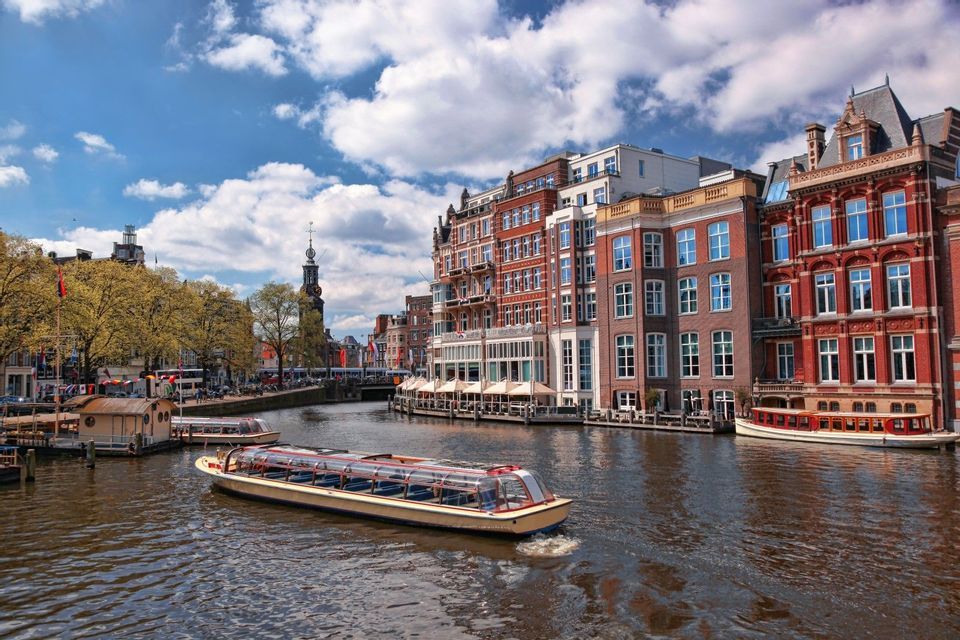 A tour boat with a glass roof cruises down a city canal lined with traditional brick buildings under a partly cloudy sky.