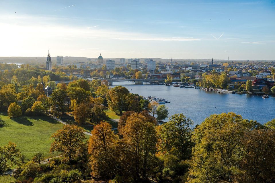 An aerial view of a park with autumn trees alongside a river, with a city skyline in the background under a clear sky.