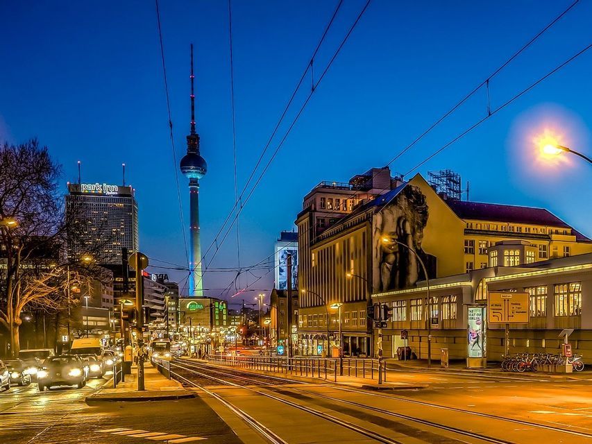 A city street at night with illuminated buildings, tram tracks, and a tall television tower against a dark blue sky.