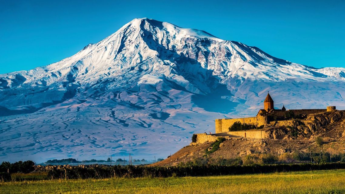 Un monastero in pietra sorge su una collina di fronte a un'imponente montagna innevata sotto un cielo azzurro e limpido.
