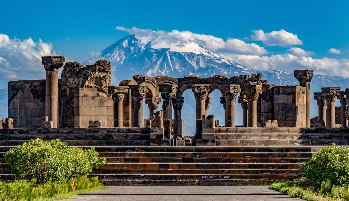 Les ruines d'un temple en pierre aux colonnes ornées se dressent devant une grande montagne enneigée sous un ciel bleu partiellement nuageux.