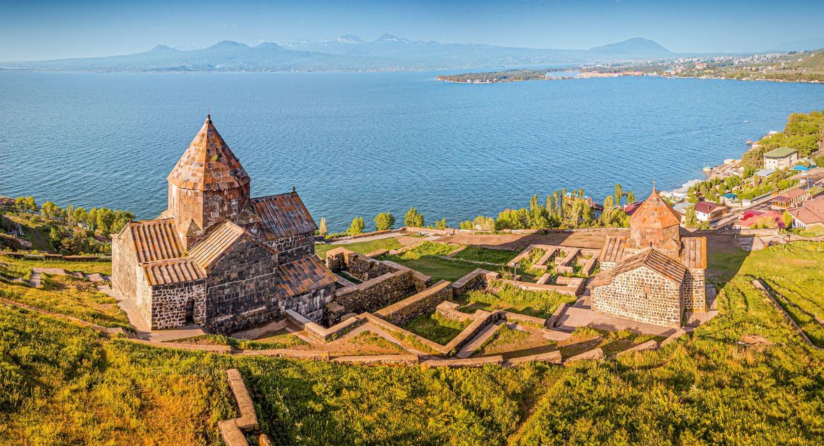 Un complexe monastique historique en pierre sur une colline verdoyante, surplombant un grand lac bleu, avec des montagnes lointaines à l'horizon sous un ciel dégagé.