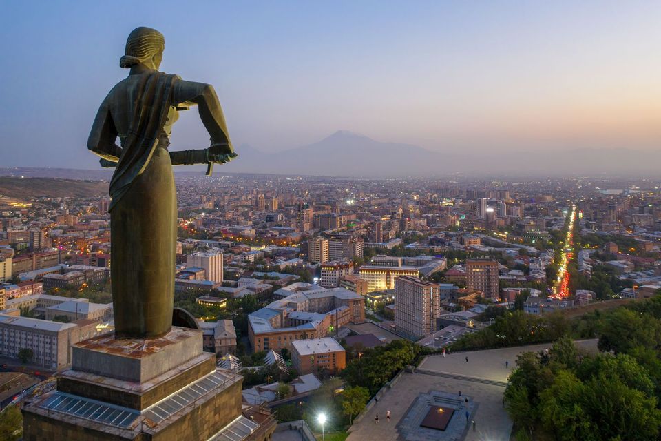 Una imponente estatua, vista de espaldas, se alza sobre una vasta ciudad iluminada al anochecer, con una montaña lejana en el horizonte.