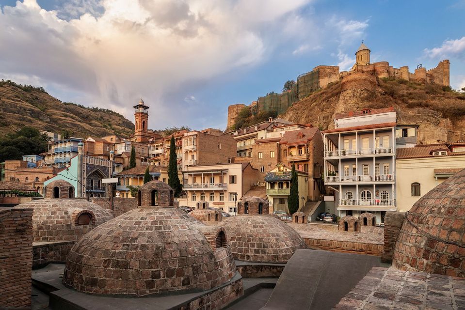 Una vista sobre cúpulas de ladrillo hacia coloridos edificios de la ciudad, anidados al pie de una gran colina coronada por una fortaleza.