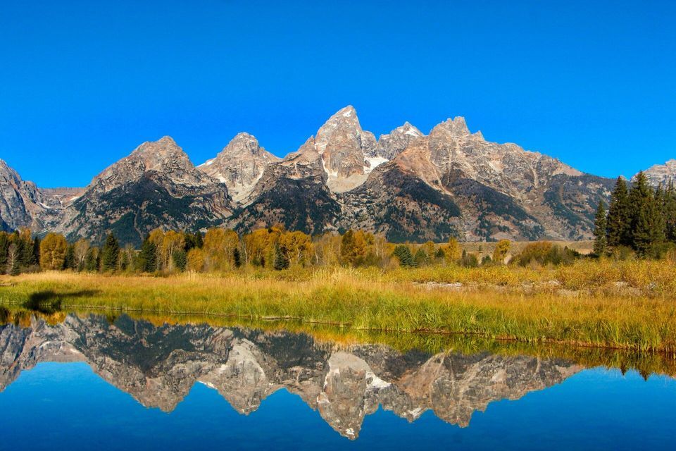 A jagged mountain range with patches of snow is reflected perfectly in a calm lake, with autumn foliage on the shore.