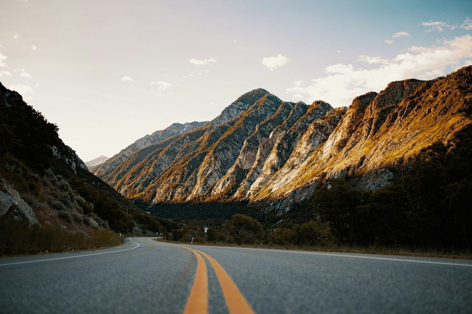 A low-angle shot of a winding asphalt road with yellow lines running through a mountain valley at golden hour.