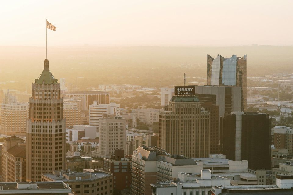 An American flag flies atop a skyscraper in a dense city skyline bathed in the hazy golden light of sunset.