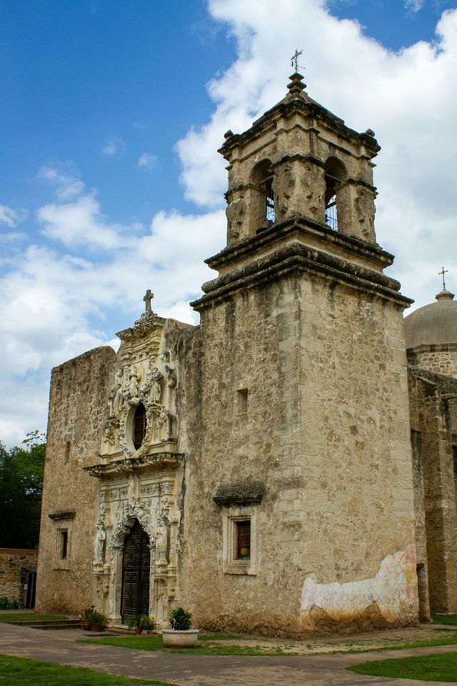 A weathered stone mission church with an ornate facade and bell tower under a partly cloudy sky.