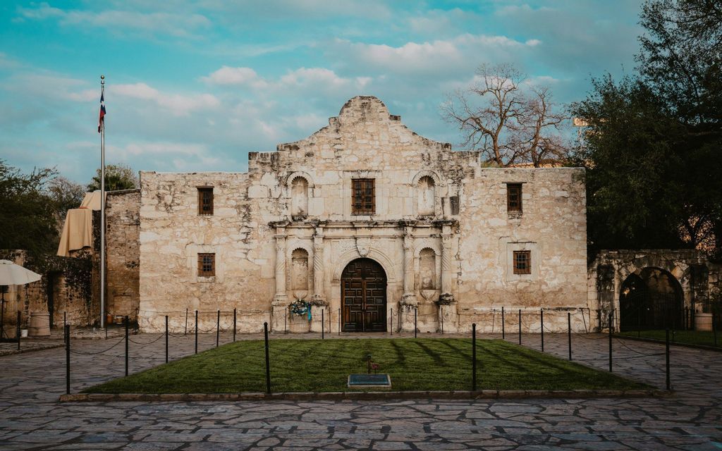 The facade of a historic stone mission building standing behind a cobblestone plaza under a cloudy blue sky.