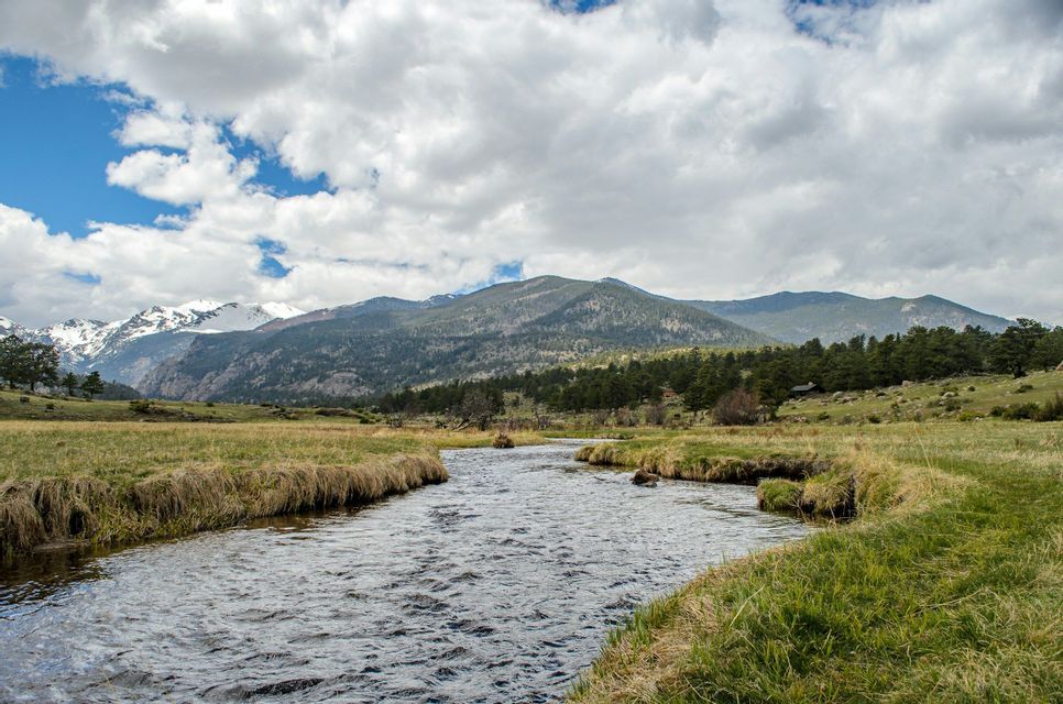 A wide river winds through a green meadow towards distant, snow-capped mountains under a partly cloudy sky.