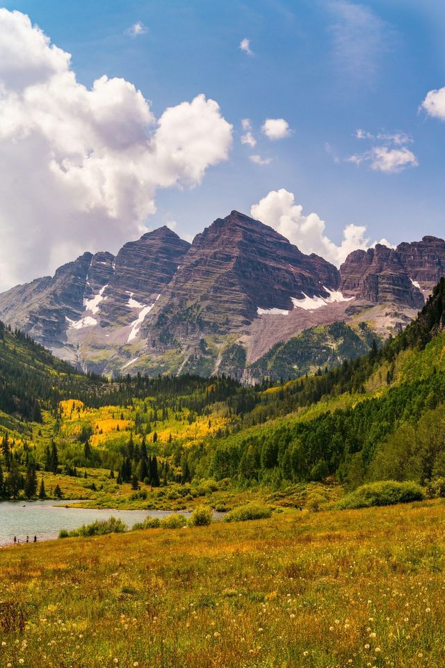 A mountain range with snow patches overlooks a valley filled with autumn-colored forests and a lake, under a partly cloudy sky.