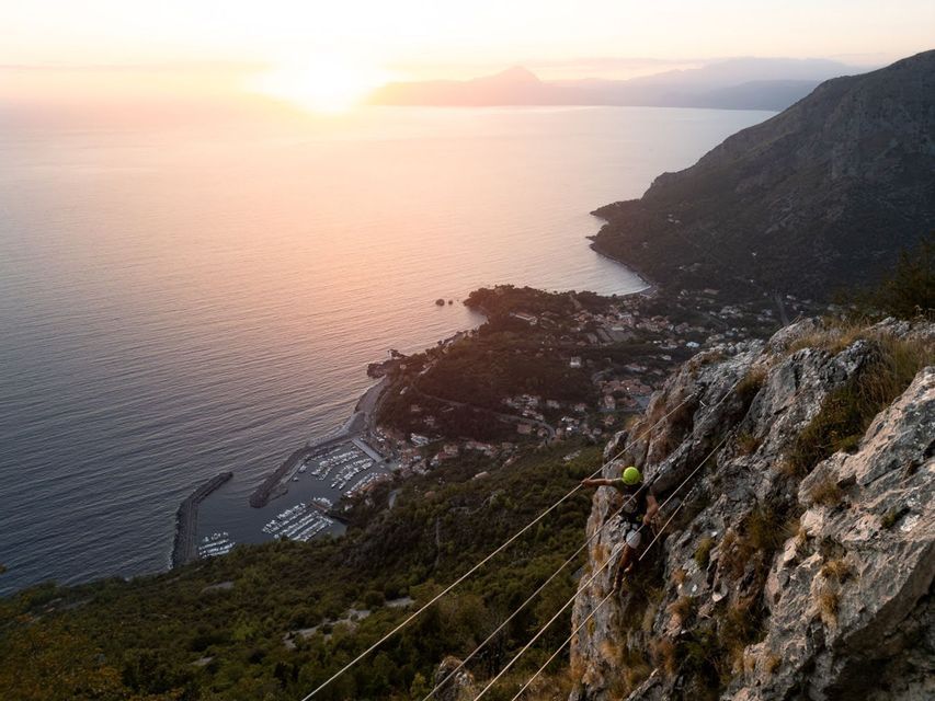 Arrampicata su scogliera, con vista aerea su marina e cittadina costiera al tramonto.