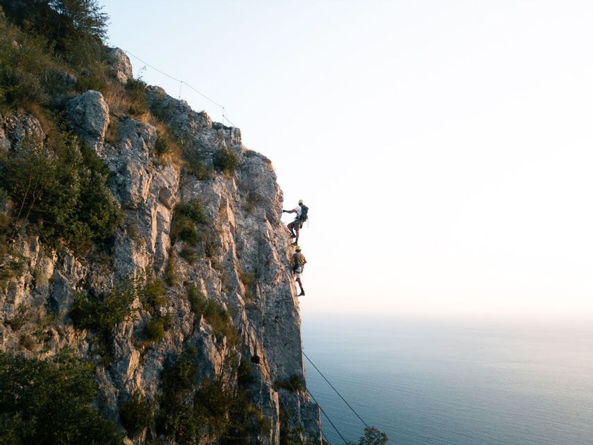 Un viaggio di gruppo WeRoad con due persone, caschi e imbracature, che praticano arrampicata su roccia su una parete a strapiombo sul mare.