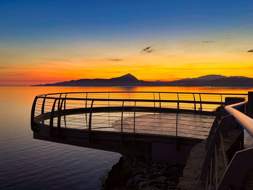 Una piattaforma di osservazione circolare si estende su un mare calmo al tramonto, con montagne lontane stagliate contro il cielo.