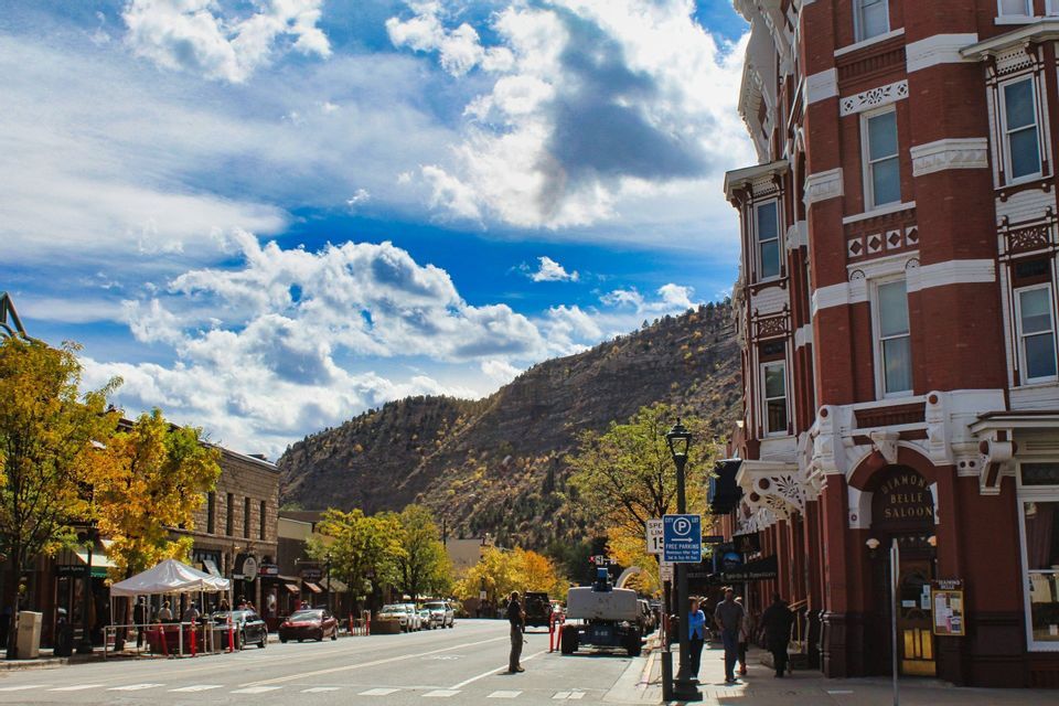 A wide street in a historic town with brick buildings and autumn trees, with a mountain rising in the background under a cloudy blue sky.