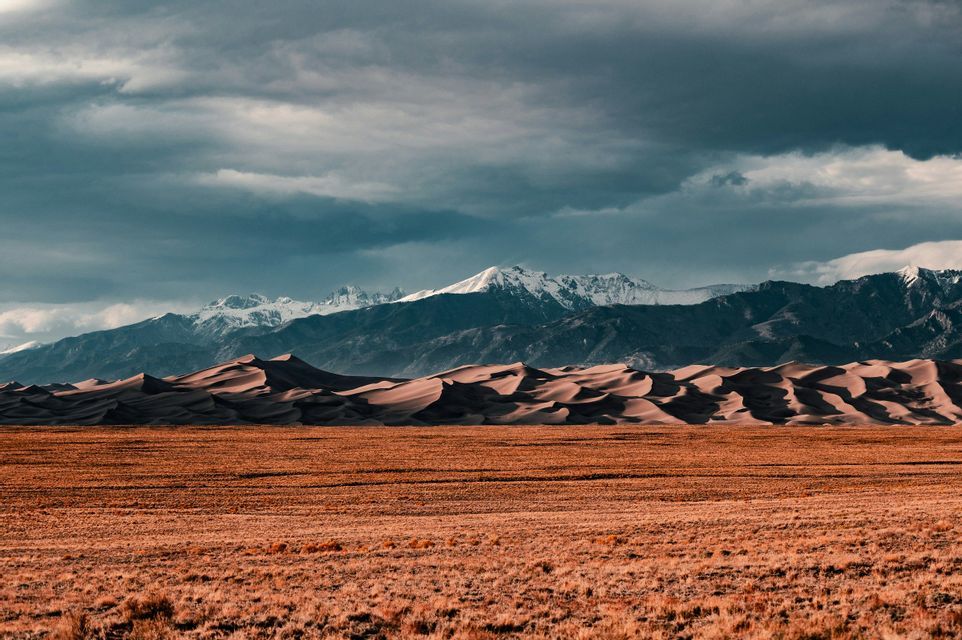 A dry, grassy plain leads to rolling sand dunes with a range of snow-capped mountains in the background under a cloudy sky.