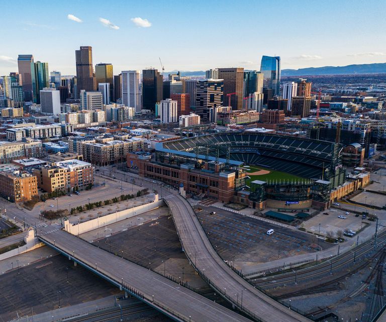 An aerial view of a large baseball stadium and empty roads with a modern city skyline and mountains in the background.