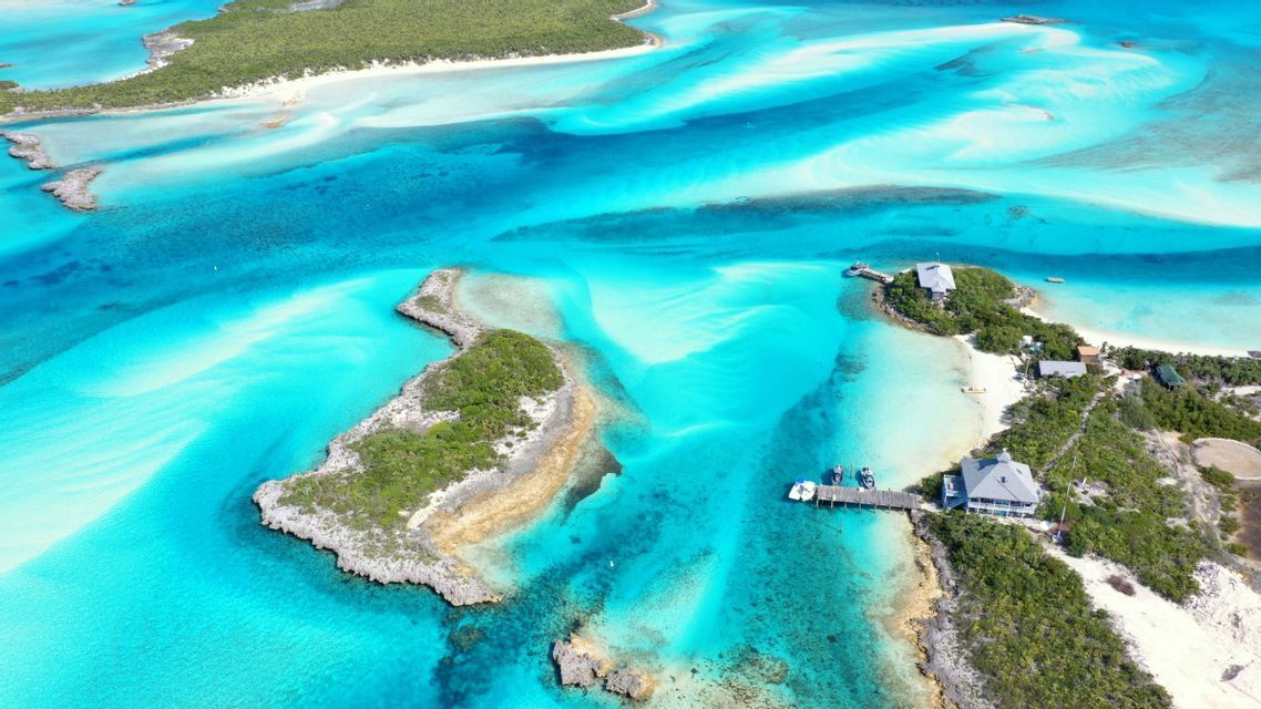 An aerial view of small, green islands and sandbars surrounded by vibrant turquoise water, with a few houses and a pier.