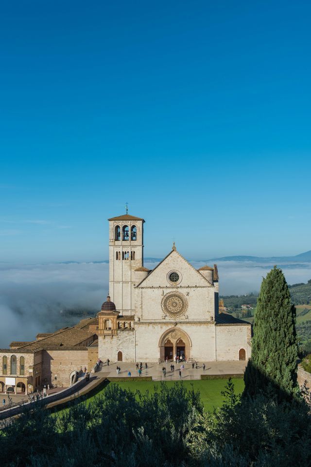 Una grande basilica in pietra con un campanile si affaccia su una valle ricoperta di nuvole basse sotto un cielo azzurro e limpido.
