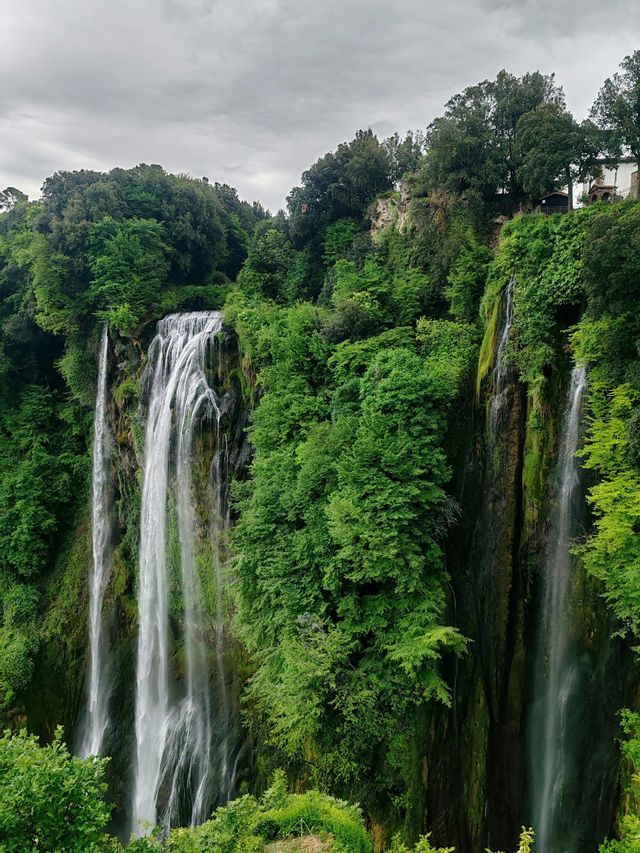 Una grande cascata scende da una scogliera coperta da fitti alberi verdi sotto un cielo nuvoloso.