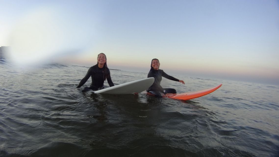 Due donne del gruppo WeRoad, in mute, sorridono sedute sulle loro tavole da surf nell'oceano.