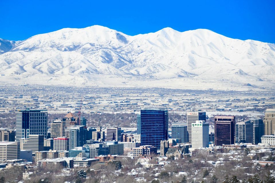 A city skyline on a clear day, with a massive snow-covered mountain range towering in the background.