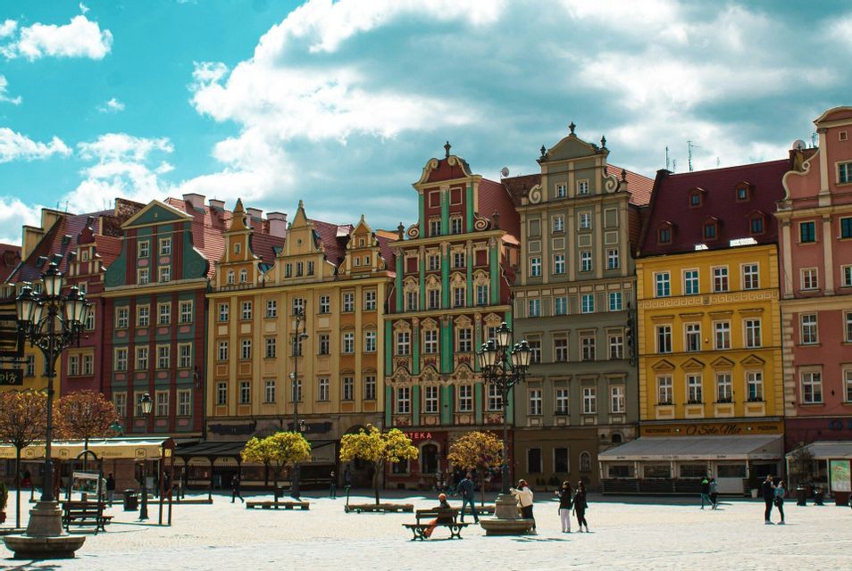 Una fila di edifici colorati e tradizionali in un'ampia piazza acciottolata dove le persone passeggiano e si rilassano sotto un cielo parzialmente nuvoloso.