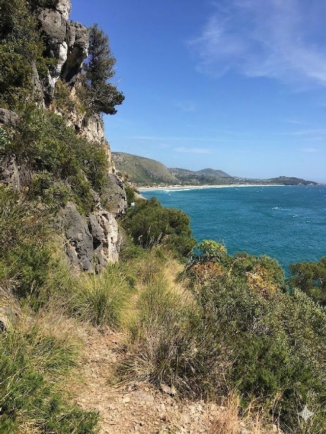 Una vista da un sentiero roccioso a picco sul mare turchese, con costa e colline in lontananza sotto un cielo azzurro.