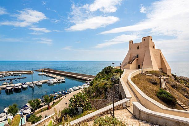 Una fortezza costiera sorge su una collina affacciata su un porto turistico pieno di barche, sotto un cielo parzialmente nuvoloso.