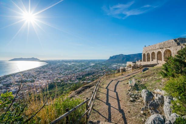 Un sentiero in cima a una collina con antiche rovine si affaccia su una città costiera e sul mare sotto un cielo limpido e soleggiato.