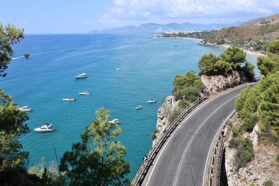 Una strada costiera tortuosa costeggia una scogliera che si affaccia su una baia turchese con diverse imbarcazioni, con un litorale distante sullo sfondo.