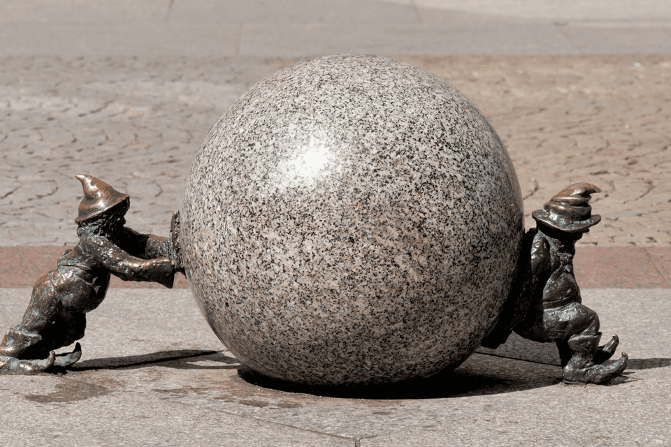 Due piccole statue di gnomi in bronzo spingono una grande sfera di granito da lati opposti su una pavimentazione in pietra.