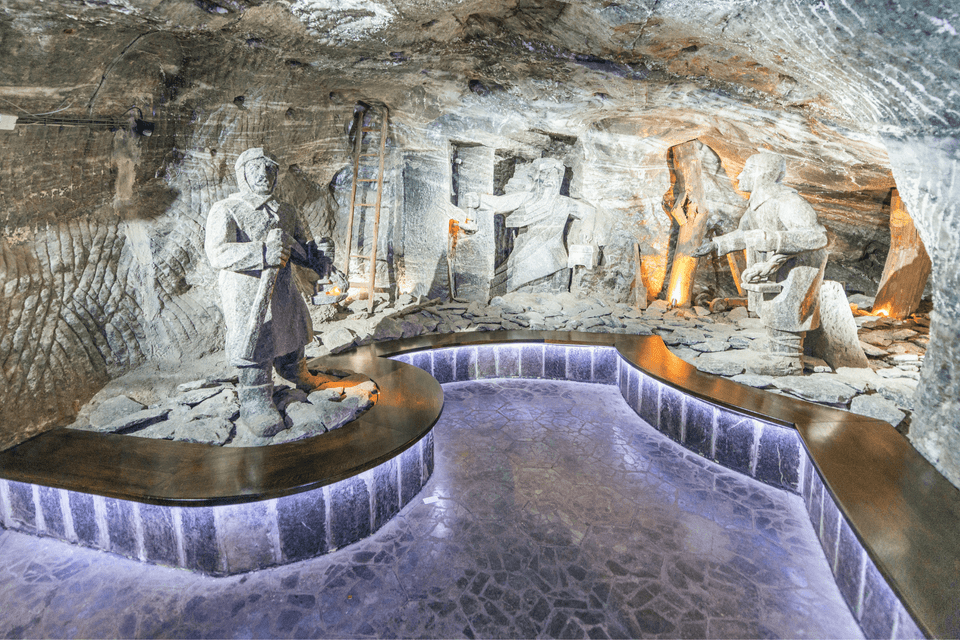 Sculture di roccia salina raffiguranti minatori sono esposte all'interno di una caverna sotterranea con una passerella illuminata e moderna.