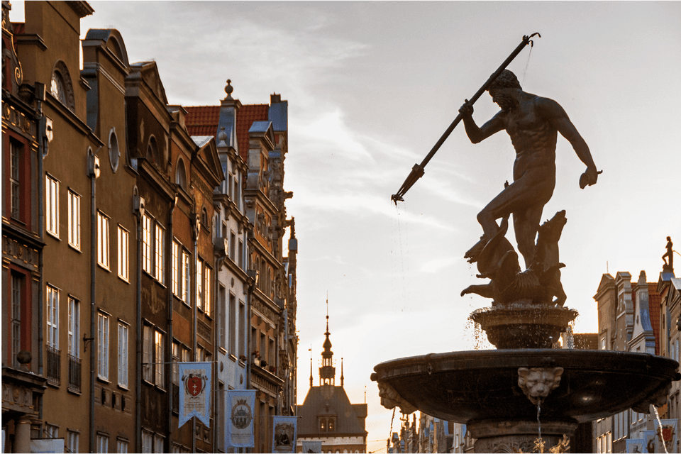 Una grande fontana in bronzo raffigurante una statua di Nettuno con un tridente, sullo sfondo di edifici storici e un cielo al tramonto.