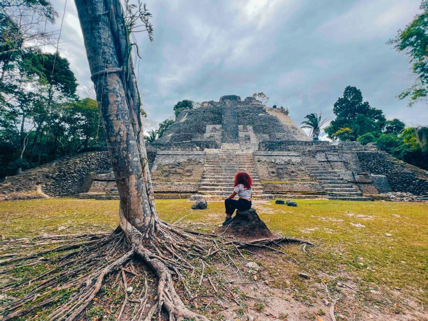 A person with red hair sits on a rock facing a large stone pyramid in a jungle clearing, with a large tree in the foreground.