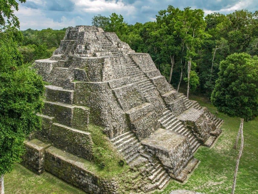 An ancient stone step pyramid rises from a clearing in a dense green jungle under a cloudy sky.
